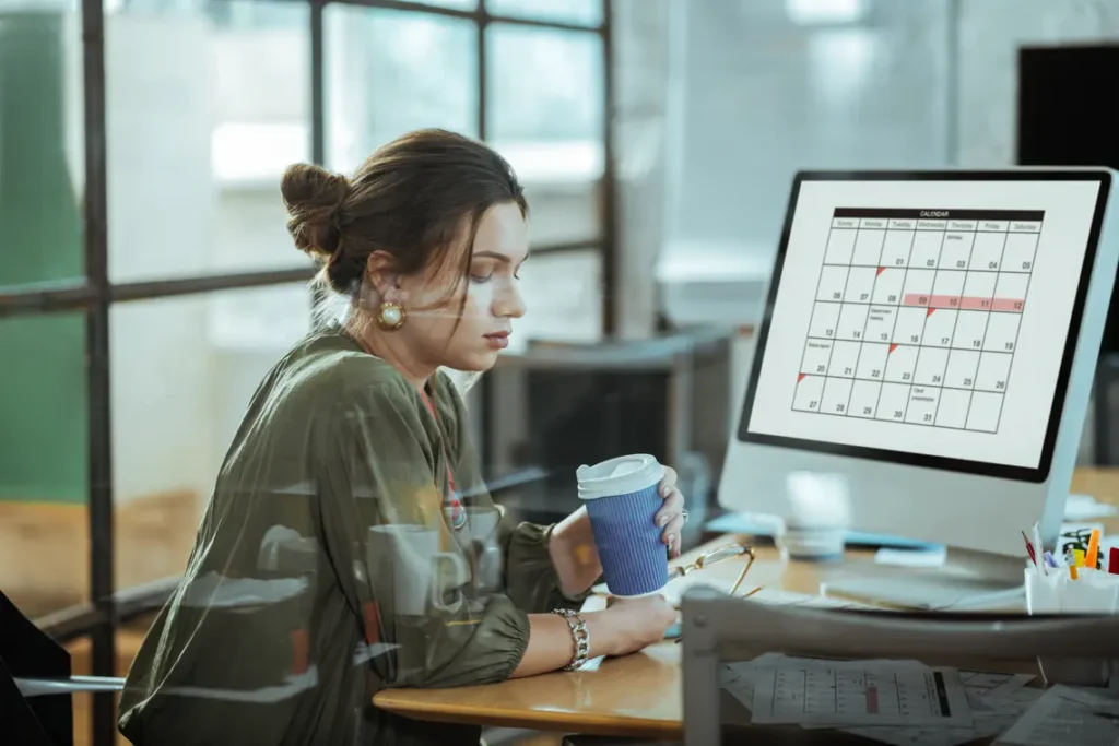 Mulher em seu ambiente de trabalho organizando sua agenda e tomando um café.